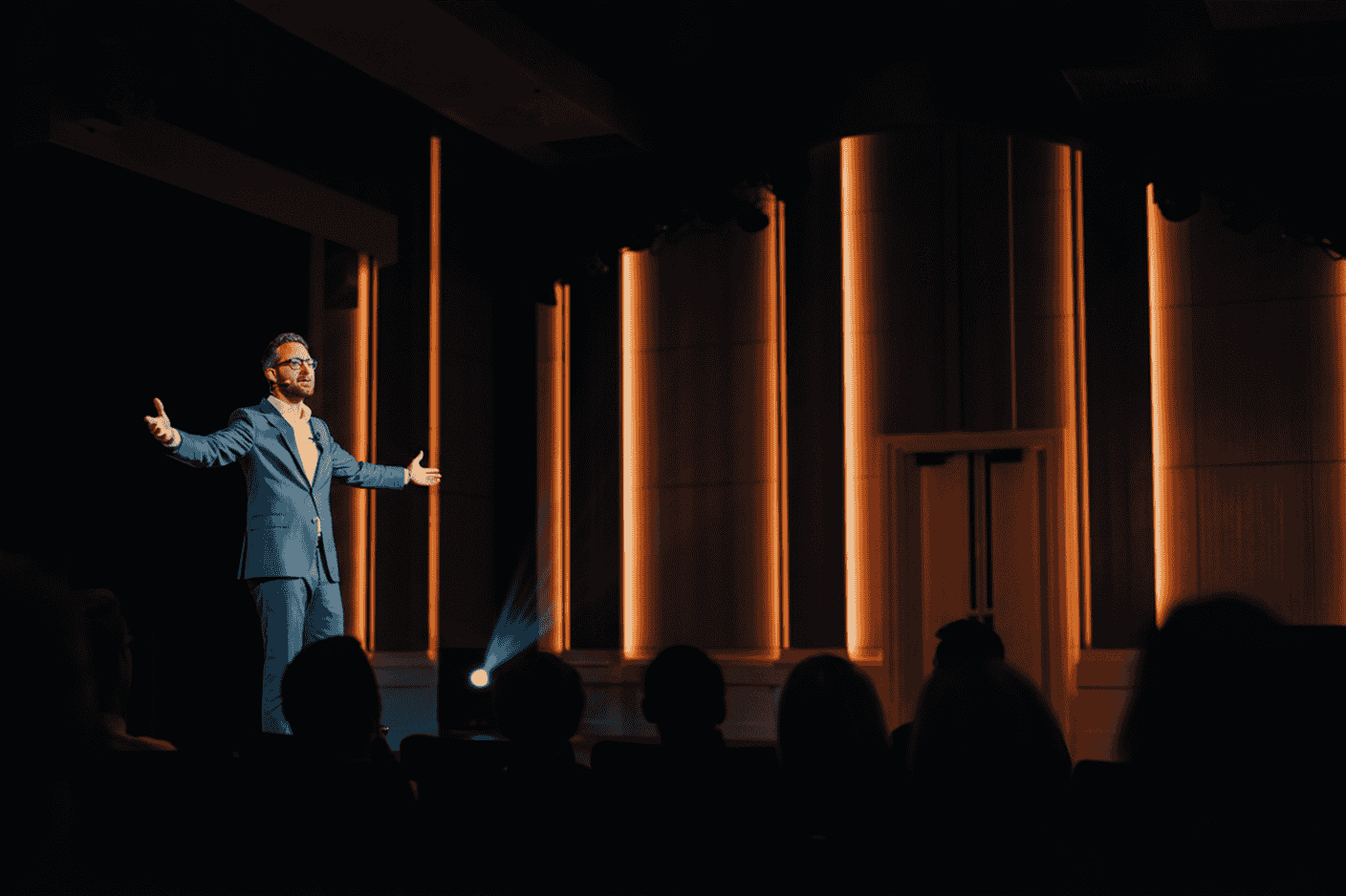a professional photograph of a confident speaker on stage with arms open in a power pose, addressing an attentive audience. The image conveys confidence, connection, and mastery with warm professional lighting and a sophisticated auditorium setting. Includes subtle visual elements that suggest transformation from anxiety to confidence