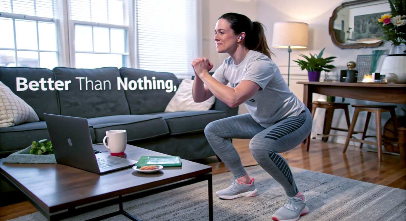 A hyper-realistic photograph of a woman in her 30s doing bodyweight squats in her living room. She is wearing comfortable loungewear (joggers and a t-shirt), not gym kit. The room is slightly messy, with a laptop on the coffee table and a mug of tea nearby, suggesting a busy day. Her expression is one of focus, simply getting a few minutes of movement in. The image captures a real, imperfect moment of choosing "better than nothing." Style: authentic, candid, and relatable, like a documentary photograph.
